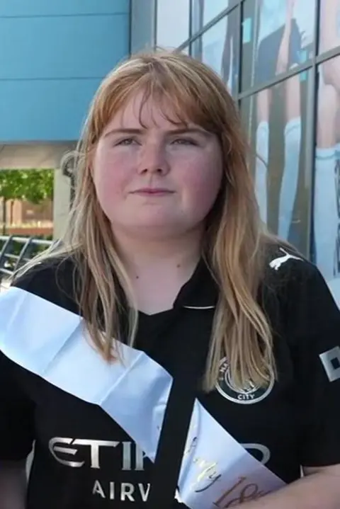 Lily has a dark Man City shirt and wears a sky blue sash. She has strawberry blonde hair. She stands in front of the Manchester City Superstore.
