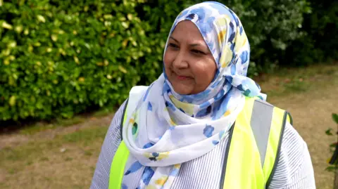 Sadia Khan, a woman wearing a headscarf and high visibility jacket. She is stood alongside a fence, with a large hedge behind her.