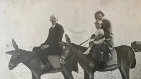 Family Photo Child in white, aged three, being held by his mother - both sitting on a donkey on Southend beach. The boy's father dressed in a dark suit and tie is astride another donkey next to them.