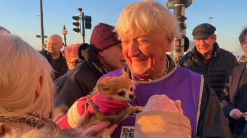 Derrick Downs holding his dog and wearing mauve bib smiling and surrounded by people in Margate at the end of his walk