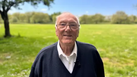 Gerry Cannell in a grassy Black Patch Park, Smethwick, wears a black jumper and white shirt and glasses.