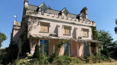 A run-down and boarded-up light-coloured building. There is overgrown foliage surrounding it and ivy crawling up the walls.