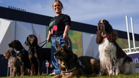 PA Media Ellie Simmonds, in black T-shirt and blue denim jeans, stands holding the leads of several dogs on a grass lawn. behind her is the wall of a building. There are five dogs next to her, three sit, one stands and the other lies on the ground.