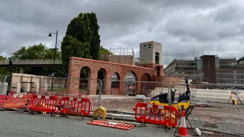 Shropshire Council A construction site shows a red brick archway structure under development with scaffolding and safety barriers surrounding it. In the background, a modern building and a pedestrian bridge are visible under a cloudy sky. 