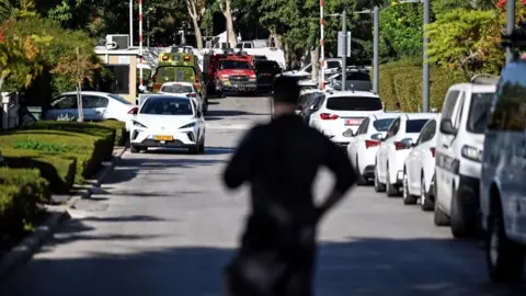 Reuters A member of Israeli security personnel stands at the entrance to a street, following a drone attack from Lebanon towards Israel amid ongoing hostilities between Hezbollah and Israel, in Caesarea, Israel, 19 October. A fire service vehicle and an ambulance can be seen in the background.