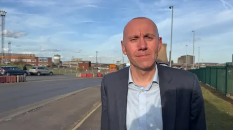 A man with a shaved head wears a blue suit with a pale blue shirt, open at the collar, in front of an industrial plant on a sunny day.