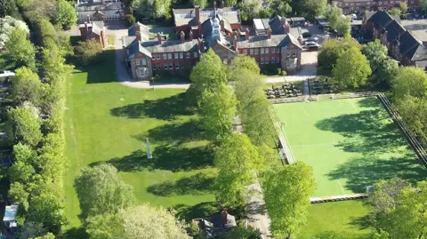 An aerial view of an ornate building with a large field in front of it as well as a playing field and trees. There are rows of houses to the left hand side of the image.