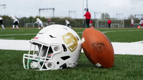 An American football and helmet placed next to each other on a pitch. People are playing American football in the background.