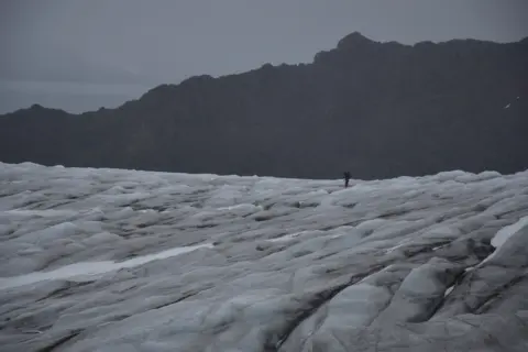 Dariusz Puczko A photography of a vast white glacier surface, with a large black mountain in the background. A person is in the middle. It gives the sense of a dangerous, foreboding place.