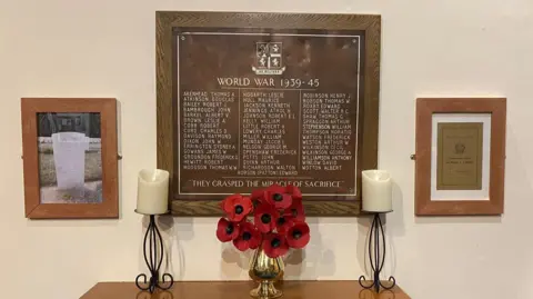 A large plaque to those who died in World War Two on the wall of a church with a large bowl of poppies in front of it 