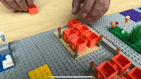 A grey Lego board with patches of grass, orange houses and tiny lights on it. A pair of hands can just be seen about to place another house on the boar.