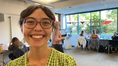 Zac Sherratt/BBC Lily Leung, student ambassador, call handler at Royal Holloway. She is smiling to camera. Lily has brown hair and glasses. She is wearing a green and yellow top. 