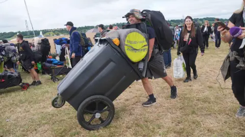 PA Media Man pushes modified wheelie bin across grass with bags and sleeping equipment in