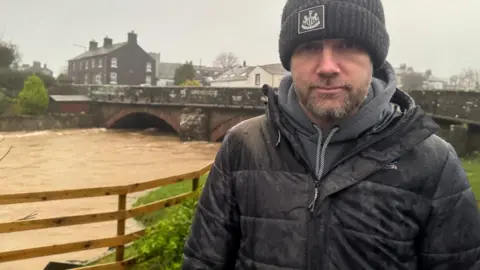 BBC Egremont town councillor David Wright who stands in front of a swollen river under an old bridge. He is wearing a black coat, black hat and grey hoodie. It is raining heavily. 