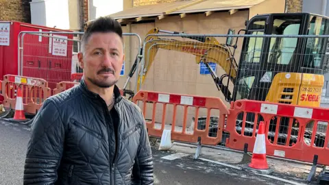 BBC Mr Marcello standing outside his home, which is fenced off with a digger outside