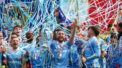 Matt Grimes is in the middle of the picture, holding the Championship Trophy in his right hand. He is surrounded by his team mates and all are being covered in streamers and confetti.
