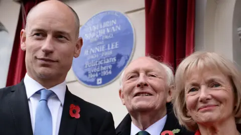 PA Media Stephen, Neil and Glenys Kinnock formally dressed wearing red remembrance poppies. Behind them is a blue circular plaque mounted on a light-coloured wall, flanked by red curtains drawn back for a ceremony.