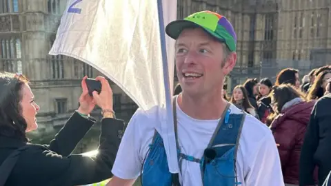 The image shows a male runner standing on a busy street near the Palace of Westminster in London. The runner is holding a large white flag attached to a pole. The runner is wearing a white T‑shirt, a colourful cap and a blue running vest with pockets and straps.