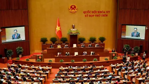 Getty Images A zoomed out shot shows rows of people sitting inside Vietnam's parliament, which is fitted out in brown and yellow shades