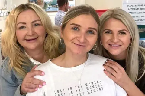 Family handout Three women smile at the camera. The woman in the centre has straight brown hair tied back, and is wearing a white top with the words “lost in the moment” written three times on it. The other two women have blond hair.