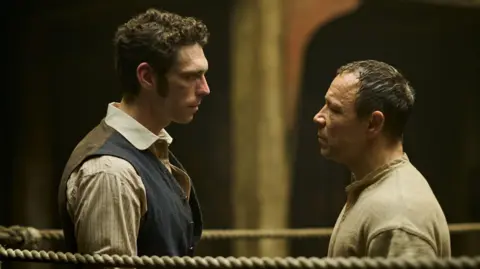Publicity picture for A Thousand Blows. It shows Nelson-Joyce (left) stood in a boxing ring facing each other. Nelson-Joyce wears a striped-shirt under a blue waistcoat and has curly dark hair. Graham wears a beige top and has short dark hair.