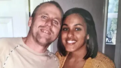 A smiling man and woman looking at the camera. The woman has dark shoulder length hair and is wearing a knitted mustard coloured top. She is stood closely next to a man, with their heads touching. He has short hair and facial stubble and is wearing a sand coloured t-shirt. 