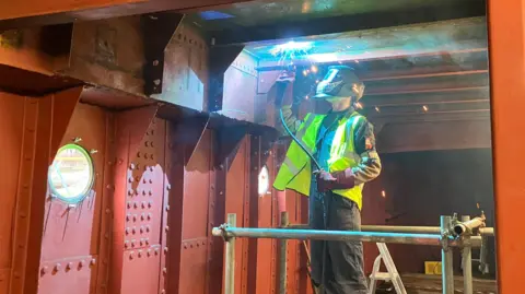 Jon Wright/BBC A person in a helmet and high-visibility jacket welding a metal ceiling inside the hull of a boat.