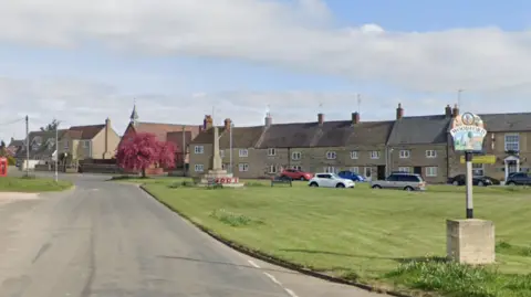 Google High Street in Woodford is mostly residential properties. There is a patch of grass to the right with a sign for Woodford and a war memorial in the background. 