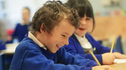 A boy wearing a blue jumper writing on a piece of paper as a girl also wearing a blue jumper watches on.