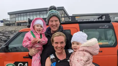 Alison Robinson Alison crouches down holding her daughter Seren on her hip. Alison has a watersports vest on and is wearing her hair in plaits. Seren, 3, is wearing a white hat and a pink floral coat with faux fur around the hood. Alison's husband Paul Robinson stands behind them wearing a blue and green hat and a black coat. He is holding daughter Rhian, 5, who is wearinga matching white hat with Seren, and a pink horse cartoon waterproof jumpsuit. The family are on a beach.