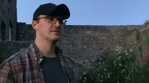 Lars Janssen stands in front of a wall at Castle Cornet. He is wearing glasses, a dark baseball cap, a dark shirt, and a plaid button-up shirt over it.