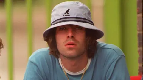 Getty Images Liam Gallagher looking slights off camera. he is wearing a white bucket hat with two thin dark stripes just about the brim pulled down to just about his eye. He has brown hair, down to the top of his blure round neck shirt.