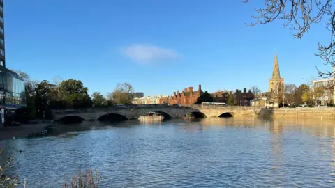 Bedford Borough Council Bedford, showing the river, the Town Bridge and a church to the left. The river level is very high. There are buildings in the distance and the sky is blue. 