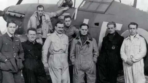 IWM/PA Media A black-and-white photo of eight men dressed in a variety of RAF uniforms and flying suits, most with slicked-back hair, posing in front of a World War Two fighter plane. One of the men is sitting on the wing, the rest are standing.