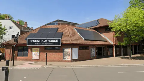 Epsom Playhouse is a building with solar panels set in the town centre. Alongside runs a road with a pedestrian crossing.