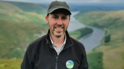 Lee Schofield Mr Swainson is wearing an "RSPB" branded black fleece and a dark green cap. He has a short brown/ grey beard and is standing in front of a rolling green landscape with a reservoir in the background.