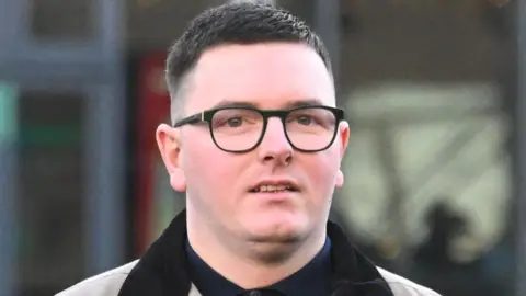 Patrick Gallagher pictured outside Belfast Crown Court. He is wearing black rimmed glasses, has short dark hair, and a grey jacket