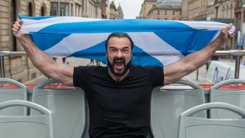 WWE Wrestler Drew McIntyre holding a saltire flag on an open top bus in Glasgow