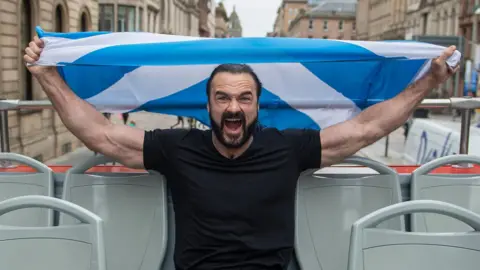Wrestler Drew McIntyre holding a saltire flag on an open top bus in Glasgow