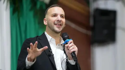 Zack Polanski, in white open necked shirt and dark jacket, addresses a Green Party rally, with microphone in hand
