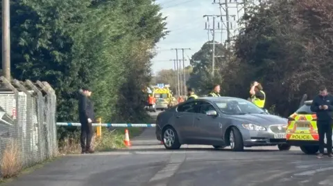 Lee Needham Police cordon on a country road. Ambulance in distance. Posh car in front of it. Police cars dotted around and officers stand around talking to each other.