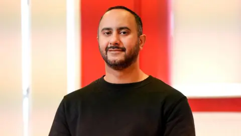 Mustafa has dark hair and has a beard and moustache. He is wearing a black sweater and is smiling at the camera. He is sat in a large hall, with red and white walls behind him.