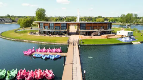 Willen Lake Willen Lake, showing a large lake, with a wooden walkway, to a small island, with a building in the middle. There are pedalos on the water and trees behind the building. There is a small white structure to the right and other buildings in the distance to the left. 