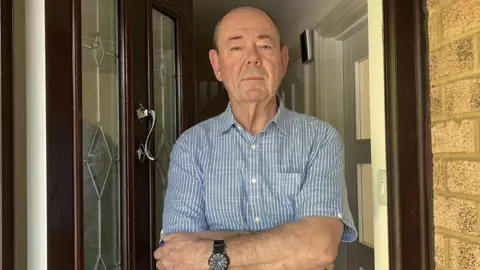 An older man wearing a blue and white striped linen shirt stands on a doorstep with his arms crossed.