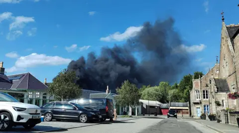 A plume of black smoke is rising into a blue sky. Cars are parked in the foreground.
