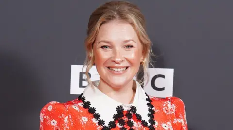 PA Media Joanna Page looking at the camera and smiling. She is wearing a red dress with a big white collar, with black flowers embroidered on it. She has blonde hair, which is tied up and gold earrings. Behind her the background is grey and there is the outline of the BBC logo in white. It is a head and shoulders shot of her. 