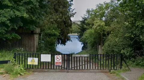 A gated gravel path leading to a large body of water surrounded by trees
