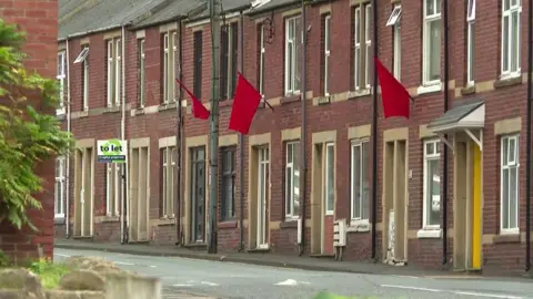 A row of terraced narrow brick houses, their entrances flush with the street. Three sport red flags on poles under the first storey windows.
