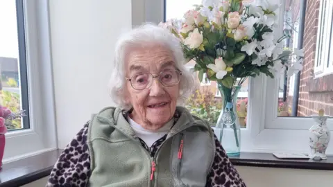 Christina Hartnell is sitting in a conservatory with a large vase of flowers behind her.