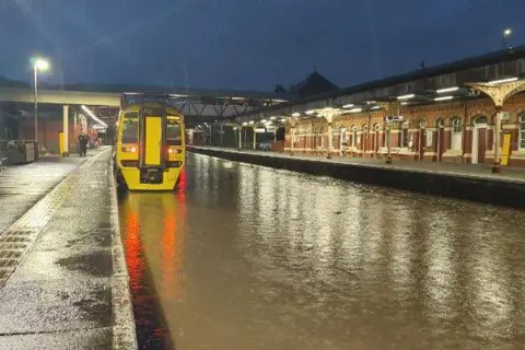 Network Rail Water flooding the railway tracks at a station. A train is parked on the line.